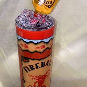Op de afbeelding: Een aangepaste Fireball Cinnamon Whisky-beker met een bijpassende fles die erin wordt gegoten. De beker is rood, oranje en zwart met het Fireball-logo en de tekst. De fles is helder met een gele vloeistof.