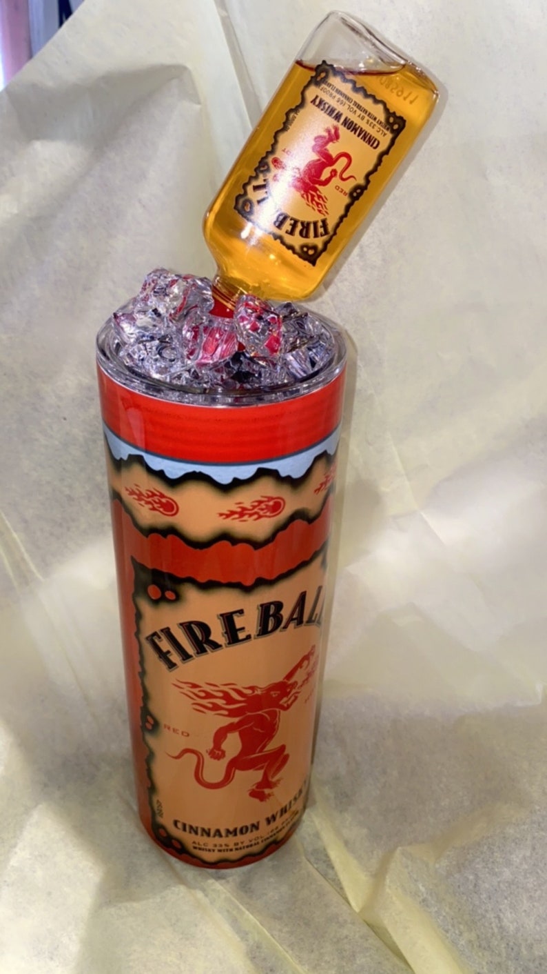 Op de afbeelding: Een aangepaste Fireball Cinnamon Whisky-beker met een bijpassende fles die erin wordt gegoten. De beker is rood, oranje en zwart met het Fireball-logo en de tekst. De fles is helder met een gele vloeistof.