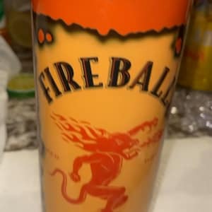 Op de afbeelding: Een cilindrische beker met een oranje gradi&euml;nt achtergrond. De beker heeft de tekst "FIREBALL" in zwarte letters en een rode duivelafbeelding. De woorden "CINNAMON WHISKY" zijn ook zichtbaar.