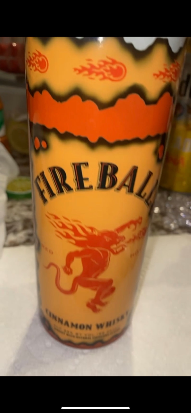 Op de afbeelding: Een cilindrische beker met een oranje gradi&euml;nt achtergrond. De beker heeft de tekst "FIREBALL" in zwarte letters en een rode duivelafbeelding. De woorden "CINNAMON WHISKY" zijn ook zichtbaar.