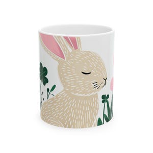 May include: White ceramic mug with a brown bunny with pink ears and closed eyes surrounded by green clover and pink flowers.