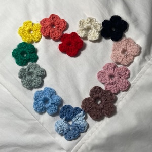 May include: A collection of colorful, crocheted flower appliques arranged in a heart shape. The flowers are in shades of yellow, orange, red, white, black, pink, green, gray, blue, and brown. The appliques are on a white background.
