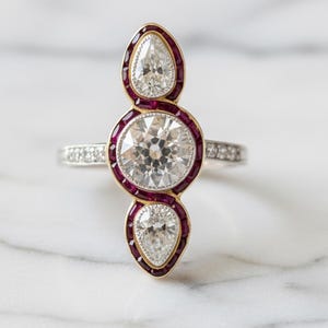 Op de afbeelding: Een elegante ring met een grote, ronde, heldere diamant in het midden, geflankeerd door twee peervormige diamanten. De diamanten zijn gezet in een gouden en robijnrode zetting, met een band versierd met kleinere diamanten. De ring wordt gepresenteerd op een wit marmeren oppervlak.