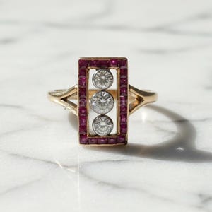 May include: A gold ring featuring a rectangular design with three clear stones and a border of small red stones. The ring has a delicate band and is set against a marble surface.