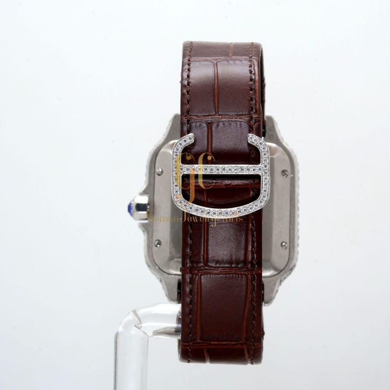 May include: A silver watch with a brown leather band and a diamond-encrusted buckle. The watch face is square and has a blue sapphire on the side.