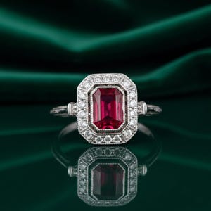 May include: A silver ring featuring a rectangular, deep red gemstone, encircled by a halo of smaller, clear stones. The ring has an ornate band and setting. The ring is displayed on a reflective surface, with a dark green backdrop.