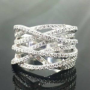 May include: A wide, silver-toned ring with a crisscross design. The ring is encrusted with numerous small, clear gemstones, creating a sparkling effect. The band is thick and wraps around the finger multiple times.