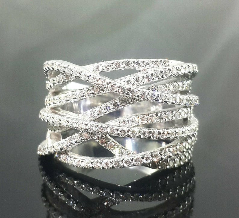 May include: A wide, silver-toned ring with a crisscross design. The ring is encrusted with numerous small, clear gemstones, creating a sparkling effect. The band is thick and wraps around the finger multiple times.