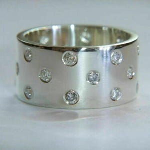 May include: A wide, silver-colored ring with multiple small, round, clear gemstones embedded in the band. The ring has a brushed metal finish and a simple, elegant design. The gemstones are evenly spaced around the ring.
