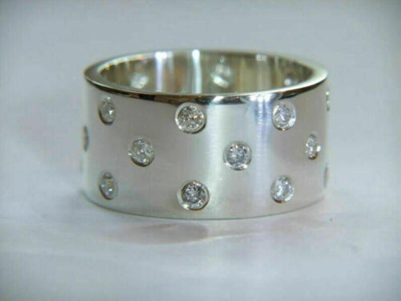 May include: A wide, silver-colored ring with multiple small, round, clear gemstones embedded in the band. The ring has a brushed metal finish and a simple, elegant design. The gemstones are evenly spaced around the ring.