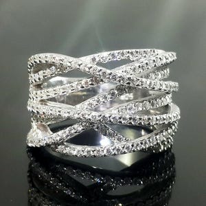 May include: A silver ring with a crisscross design, encrusted with numerous small, sparkling stones. The ring's intricate pattern creates a layered effect, reflecting light and adding to its brilliance. The ring is displayed on a reflective surface.