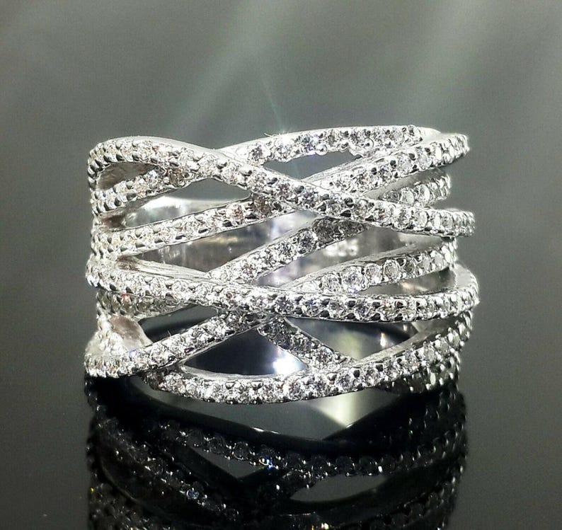 May include: A silver ring with a crisscross design, encrusted with numerous small, sparkling stones. The ring's intricate pattern creates a layered effect, reflecting light and adding to its brilliance. The ring is displayed on a reflective surface.