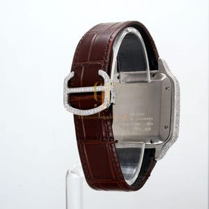 May include: A silver and diamond Cartier watch with a brown leather band. The watch face is surrounded by diamonds and the band has a diamond buckle. The watch face has the text "Swiss Made" and "Cartier" on it. The watch is water resistant to 100 meters. The watch is made of stainless steel.