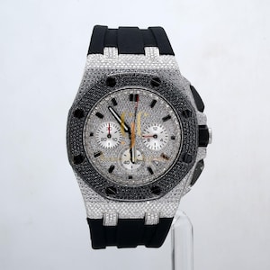 May include: A luxury wristwatch with a silver face and a black rubber strap. The watch is adorned with small, sparkling stones around the bezel and face. It features three sub-dials and a date display. The hands are gold-coloured.