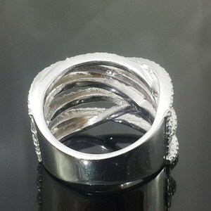 May include: A silver-colored ring with a multi-band design. The ring features multiple interwoven bands, some of which are encrusted with small, sparkling stones. The ring has a polished, reflective surface, and the design creates a layered effect.