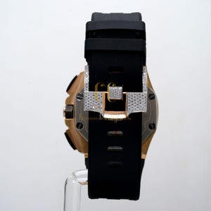 May include: A gold and black wristwatch with a diamond-encrusted clasp. The watch has a black rubber strap and a round face.