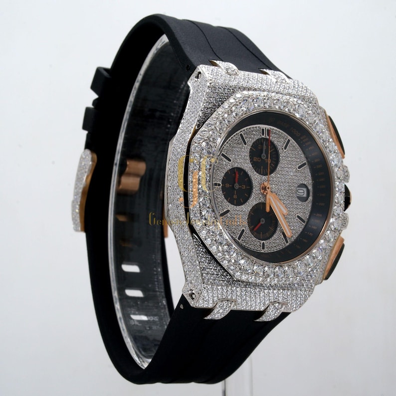 May include: A diamond-encrusted wristwatch with a black rubber band and a rose gold bezel. The watch face has a chronograph with three sub-dials.