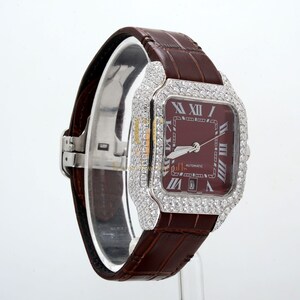 May include: A white gold wristwatch with a brown leather band. The watch face is square and has a red dial with Roman numeral hour markers. The watch is set with diamonds around the bezel.