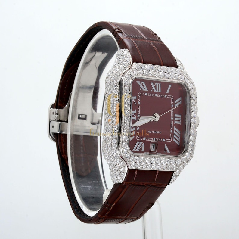 May include: A white gold wristwatch with a brown leather band. The watch face is square and has a red dial with Roman numeral hour markers. The watch is set with diamonds around the bezel.