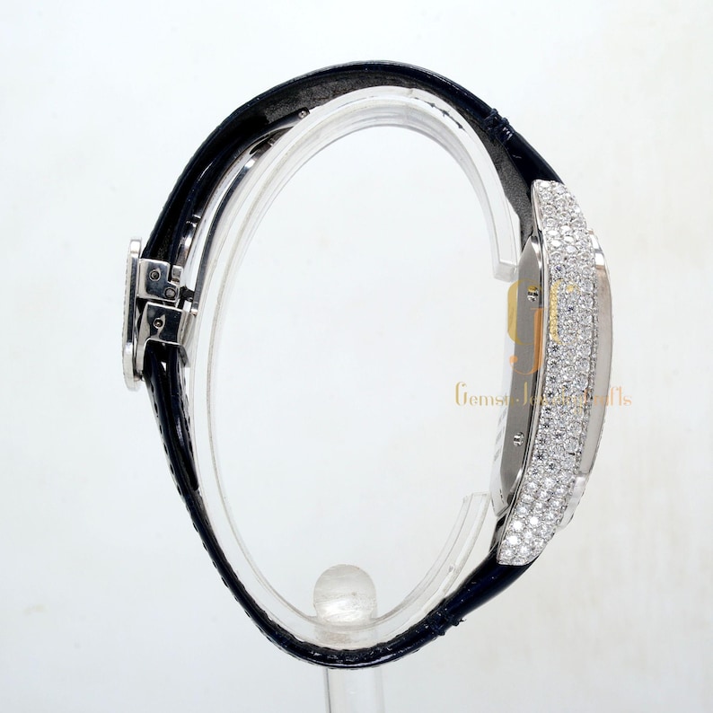 May include: A silver watch with a black leather band. The watch face is covered in diamonds.
