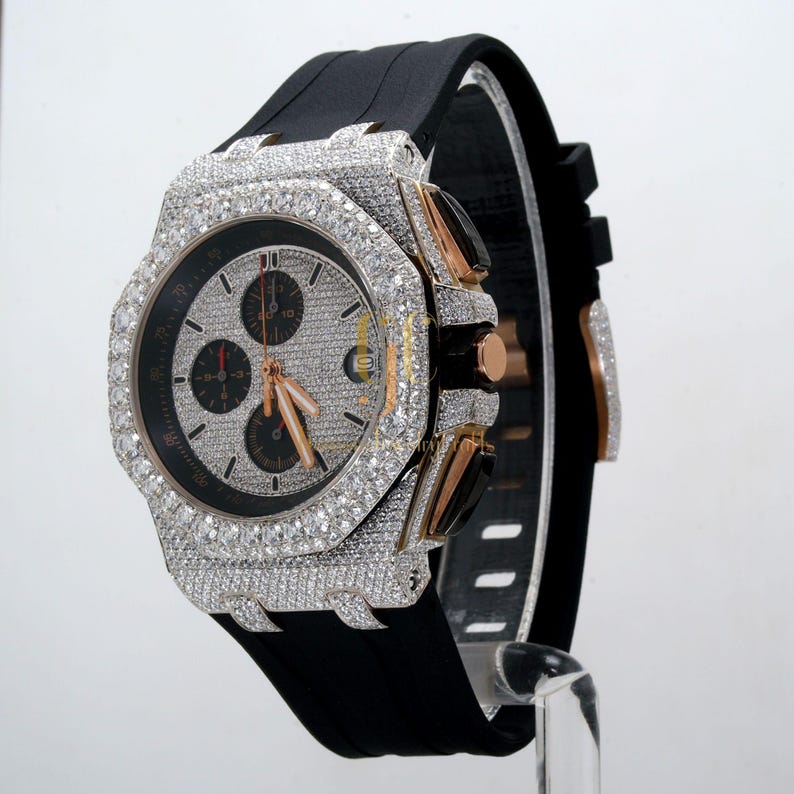 May include: A black rubber band watch with a silver and rose gold case and a diamond-encrusted bezel. The watch has a chronograph function and a white dial with black hands and markers.