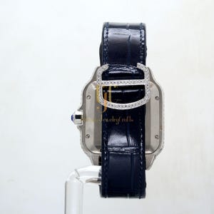 May include: A silver watch with a diamond-encrusted bezel and a blue leather strap. The watch has a square face and a rectangular case.