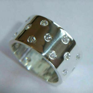 May include: A wide, silver-colored ring with a polished finish. The ring is adorned with multiple small, round, clear gemstones evenly spaced across the surface, creating a sparkling effect. The ring's design is simple yet elegant.