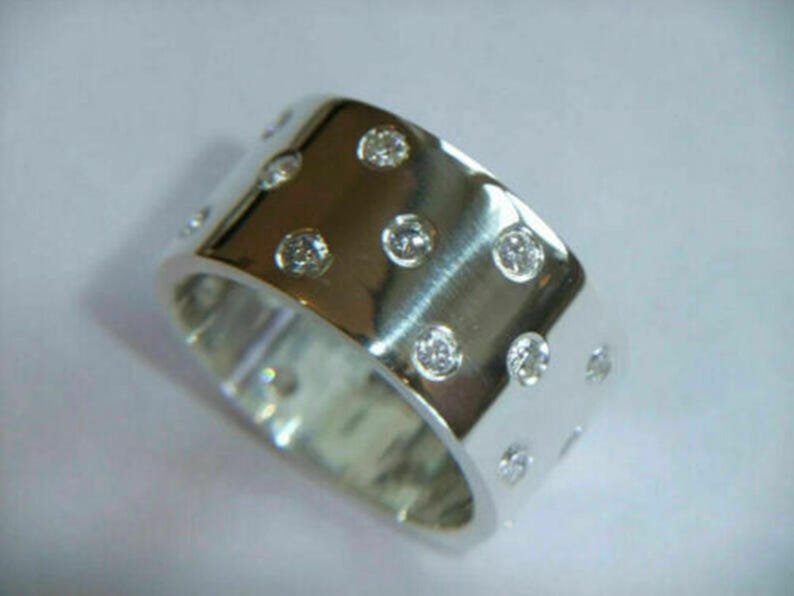 May include: A wide, silver-colored ring with a polished finish. The ring is adorned with multiple small, round, clear gemstones evenly spaced across the surface, creating a sparkling effect. The ring's design is simple yet elegant.