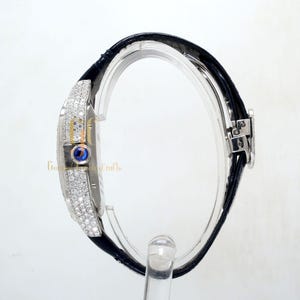 May include: A silver wristwatch with a black leather band and a diamond-encrusted bezel. The watch face has a blue sapphire cabochon at the 12 o'clock position.