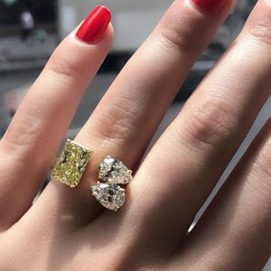 May include: A close-up of a ring featuring a large, rectangular yellow diamond and two pear-shaped diamonds. The ring is set on a finger with red nail polish.