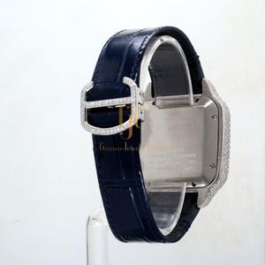 May include: A silver Cartier watch with a diamond-encrusted bezel and a blue leather strap. The watch face is silver and has a date window. The watch is water resistant to 30 meters. The watch is made in Switzerland.