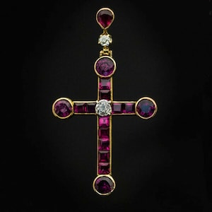 Puede incluir: Un colgante de oro en forma de cruz, adornado con piedras de rubí y diamante. La cruz está engastada con rubíes de forma cuadrada y un diamante redondo más grande en el centro.