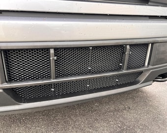 Könnte beinhalten: Nahaufnahme der hinteren Stoßstange eines Fahrzeugs mit einem schwarzen Maschengitter. Das Gitter wird von einer dunkelgrauen Metallstruktur mit sichtbaren Befestigungselementen umrahmt. Die Stoßstange ist hellgrau.