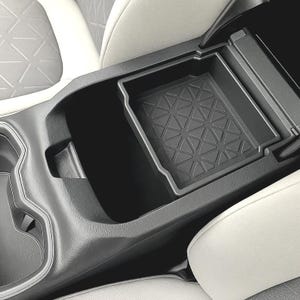 Puede incluir: Una bandeja negra para la consola del coche con un patrón geométrico de triángulos. La bandeja está diseñada para encajar en la consola central de un coche. El interior del coche tiene asientos blancos y grises.
