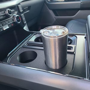 May include: A stainless steel tumbler with a clear lid is in a car's centre console cup holder. The cup holder is black and has a second, empty cup holder. The car's interior is visible.