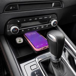 May include: Car interior showing a smartphone in a pink case, displaying 3:39 on the screen. A white USB charger is plugged in. Climate control buttons and a gear shift are also visible.