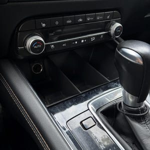 Peut inclure: Intérieur de voiture noir avec des boutons de climatisation, un levier de vitesses et des compartiments de rangement. Le panneau de commande de la climatisation comprend un bouton "AUTO" et une molette. Le levier de vitesses est dans la console centrale.