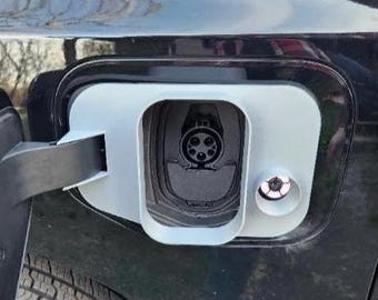 Könnte beinhalten: Nahaufnahme eines Ladeanschlusses für Elektrofahrzeuge. Der Anschluss ist schwarz und hat eine weiße Umrandung. Die Ladeanschlussklappe ist geöffnet und gibt die Ladebuchse frei. Ein kleines, kreisförmiges Schloss ist sichtbar.