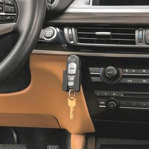 May include: A black car key holder with a key and key fob attached, mounted on a car's dashboard. The key is gold-coloured, and the interior of the car is tan and black.