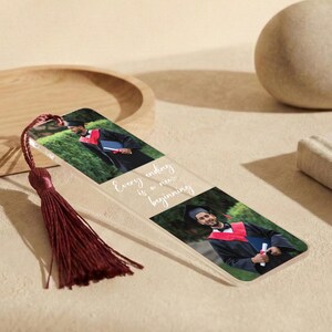 May include: A clear acrylic bookmark with a burgundy tassel. The bookmark features two photos of a person in a graduation gown and the text "Every ending is a new beginning". The bookmark is on a light-colored surface.