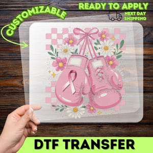 May include: A clear DTF transfer sheet featuring a graphic of two pink boxing gloves tied together with a pink ribbon. The design includes a pink checkered background, white daisies, and small pink flowers. The text CUSTOMIZABLE and NEXT DAY SHIPPING is visible.