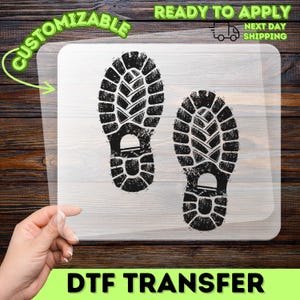May include: A clear DTF transfer sheet featuring two black, distressed boot print designs. The graphic displays the textured tread patterns of footwear soles. The sheet is transparent, allowing the underlying surface to be visible through the material.