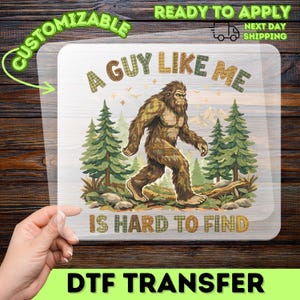 May include: A clear DTF transfer sheet featuring a graphic of a brown Bigfoot walking through a forest of green pine trees with mountains in the background. The text A GUY LIKE ME IS HARD TO FIND is printed in a plaid-patterned font above and below.