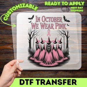 May include: A clear DTF transfer sheet featuring a graphic of five figures in pink dresses and pointed hats standing in a forest. The text reads In October We Wear Pink with a pink ribbon symbol above the figures and black bats flying overhead.
