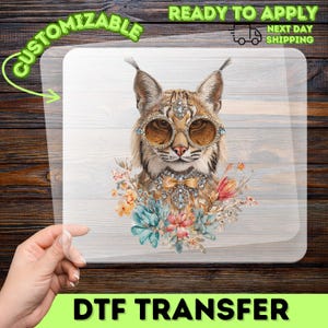 May include: A clear DTF transfer sheet featuring a graphic illustration of a lynx wearing round, jewel-encrusted glasses and a matching jeweled collar with a bow, surrounded by a cluster of colorful flowers in shades of blue, pink, and orange.