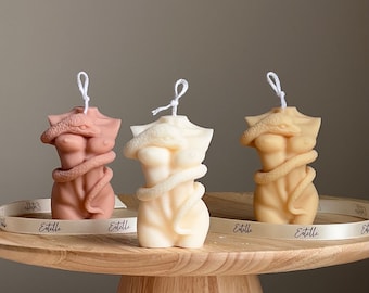 May include: Three decorative candles shaped like a human torso with a snake wrapped around it. The candles are in three different colors: brown, beige, and white. The candles are on a wooden pedestal.