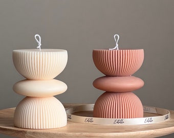 May include: Two decorative candles with a ribbed design. The candle on the left is white and the candle on the right is a light pink color. Both candles are stacked in a three-tiered design. The candles are sitting on a wooden table with a white ribbon that says "Etelle" on it.