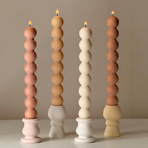 May include: Four pillar candles with a unique design. Each candle has a series of spherical shapes stacked on top of each other. The candles are in shades of pink, tan, white, and brown. Each candle is lit with a small flame.