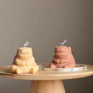 May include: Two hand-poured candles shaped like cats, one is beige and the other is brown. Both candles have a white wick and are sitting on a wooden pedestal with a white ribbon that says "Etelle" on it.