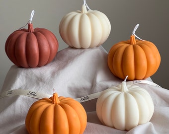 May include: Five decorative pumpkin-shaped candles in various colors, including white, brown, and orange. The candles are arranged on a white fabric surface.